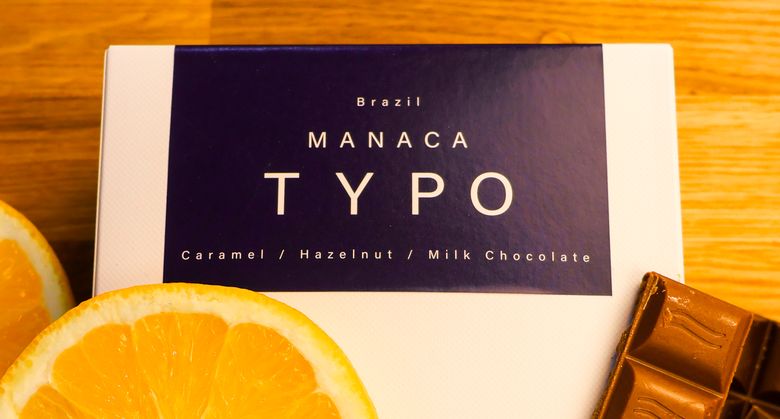 Typo Manaca coffee box from Brazil, caramel, hazelnut, milk chocolate, with half an orange and a bar of milk chocolate on a wooden table
